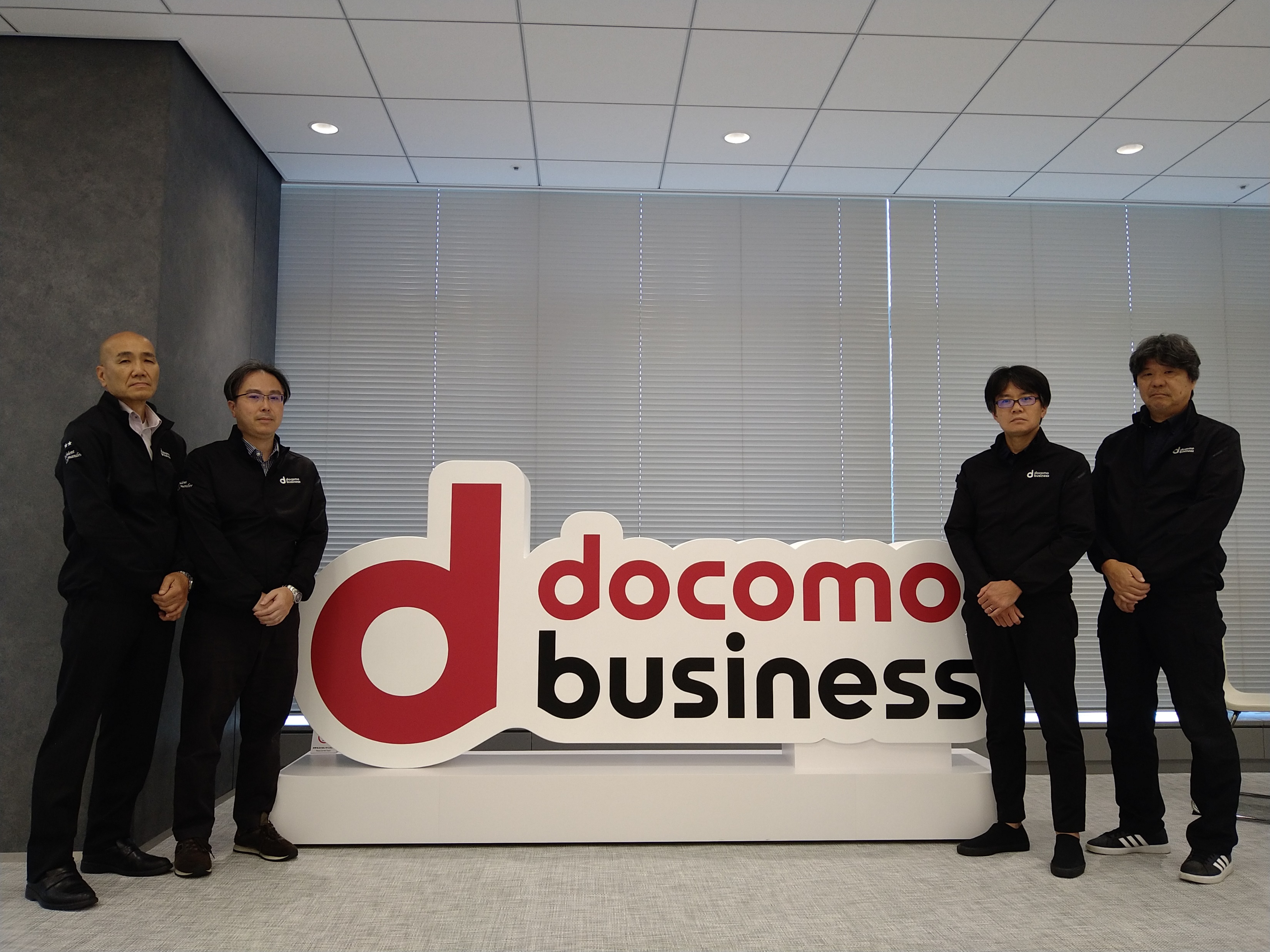 NTTドコモビジネス株式会社のロゴを背景にして、社員4名が撮影された写真